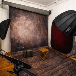 Fotografia Paweł Wojtaszek - studio fotograficzne Poznań - Studio fotograficzne z tłem w odcieniach brązu, drewnianą podłogą, walizką, stołkiem oraz sprzętem oświetleniowym, w tym softboxem GlareOne Hexa 120. Widoczny również reflektor i statywy.
