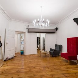 Fotografia Paweł Wojtaszek - studio fotograficzne Poznań - Studio fotograficzne z lampą błyskową, papierowymi kwiatami na ścianie, czerwonym siedziskiem z wysokim oparciem, szarym fotelem, wózkiem wiklinowym dla lalek, drewnianą podłogą i otwartymi...