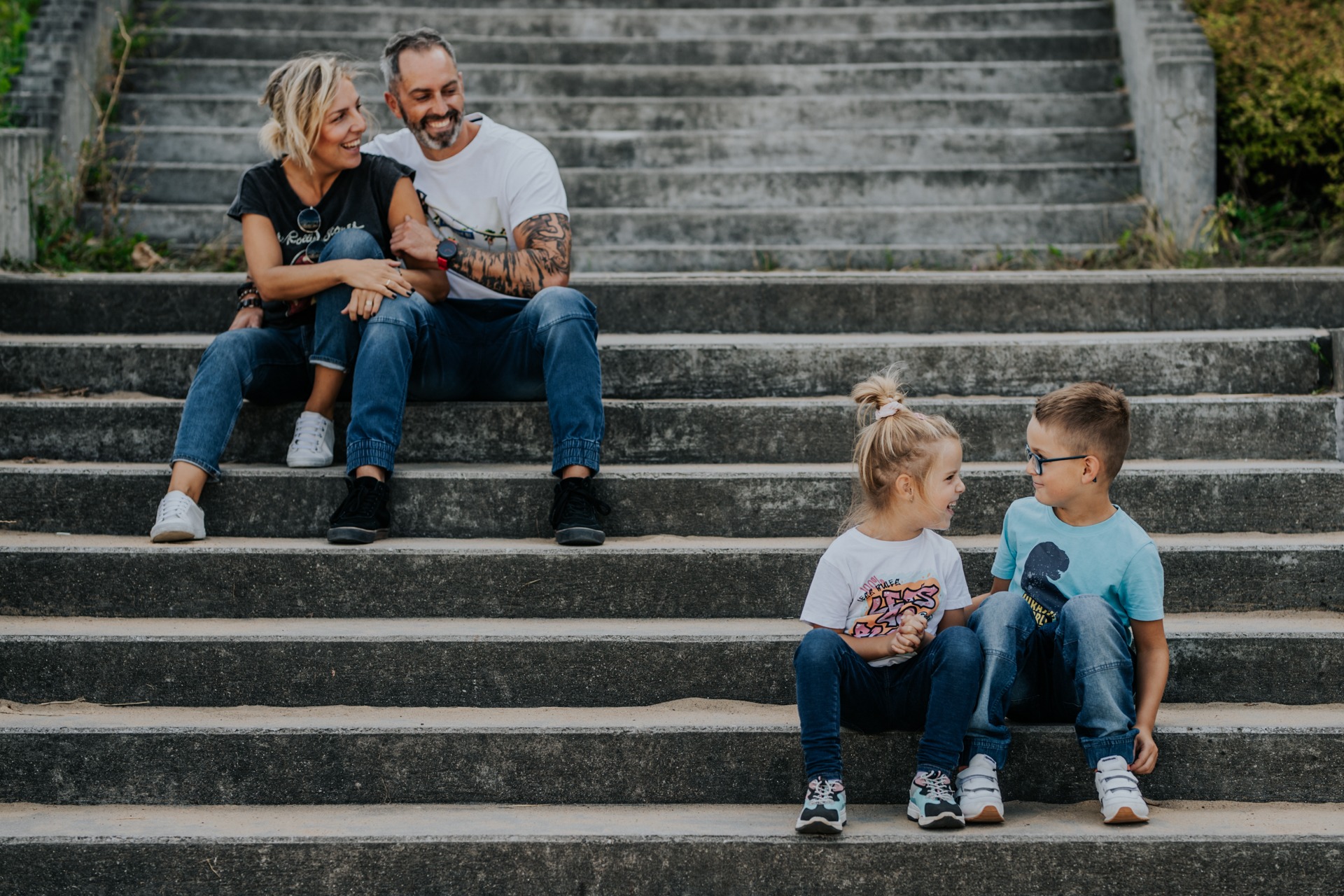 Uśmiechnięta rodzina z dwójką dzieci siedzi na betonowych schodach; tata z tatuażami na ramionach, mama w okularach na koszulce, dziewczynka z włosami spiętymi w kok, chłopiec w okularach.