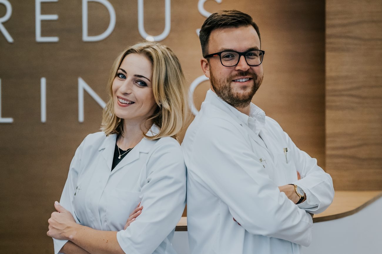 Portret dwojga uśmiechniętych lekarzy w białych fartuchach, stojących plecami do siebie na tle ściany z napisem 'REDUS CLINIC'.
