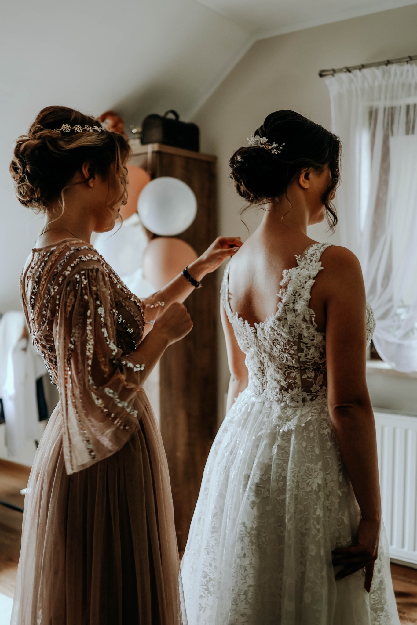Panna młoda w koronkowej sukni ślubnej, pomagająca jej druhna w sukni z cekinami, przygotowania do ślubu w pokoju z białymi zasłonami i balonami w tle.