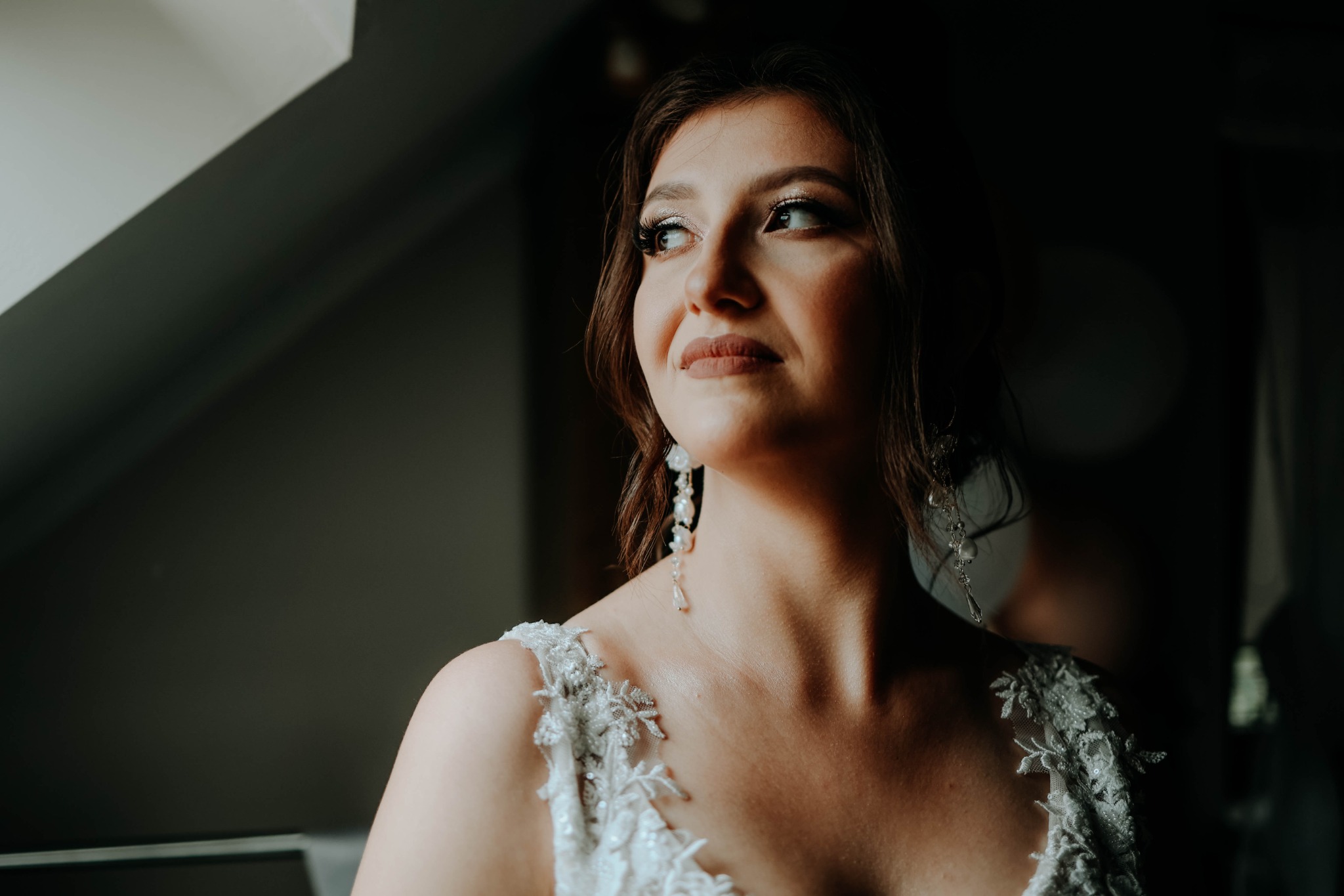 Panna młoda w białej sukni z koronkowymi aplikacjami, patrząca w lewo w kierunku okna, z delikatnym makijażem i perłowymi kolczykami.