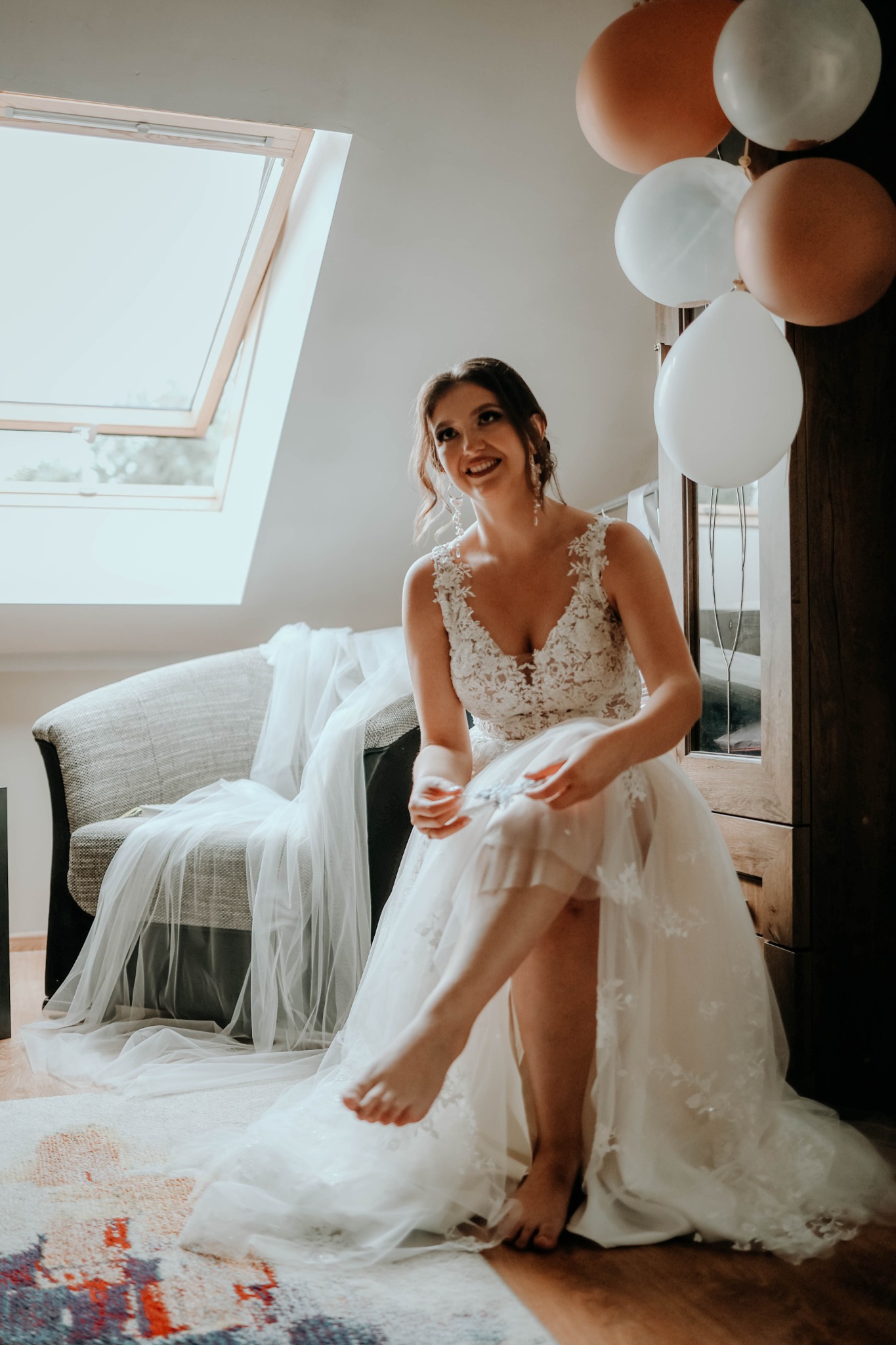 Uśmiechnięta panna młoda w koronkowej sukni siedzi na fotelu przy oknie poddaszowym, zakładając podwiązkę na nagą nogę, w tle balony i drewniana szafa.