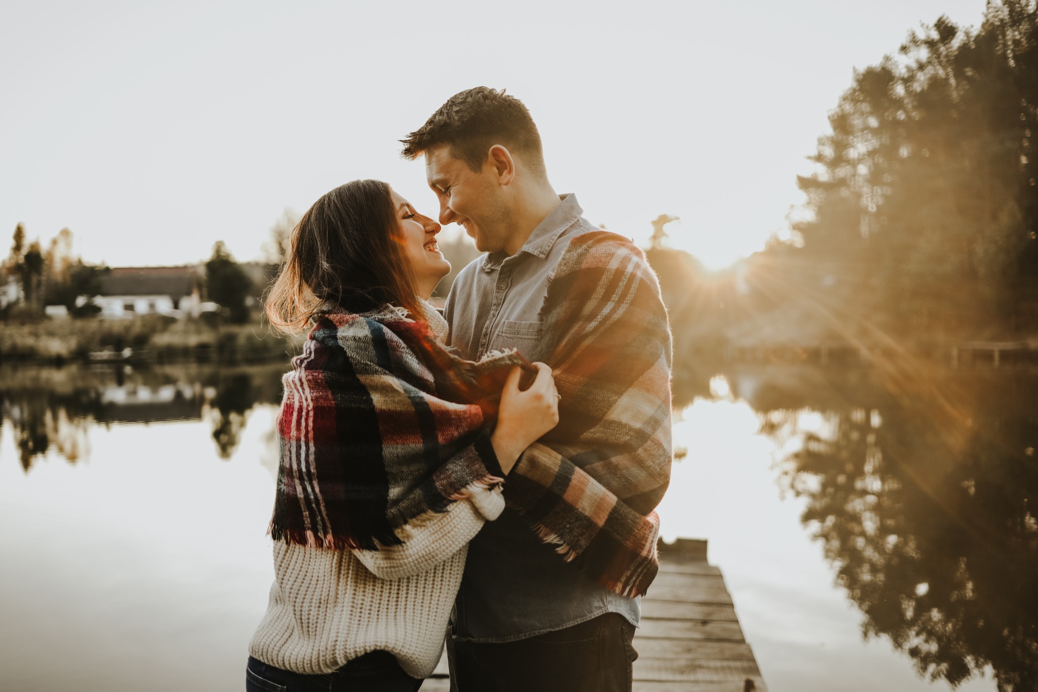 Zakochana para otulona kocem w kratę, stojąca na drewnianym pomoście nad jeziorem, twarzami blisko siebie w ciepłym świetle zachodzącego słońca.