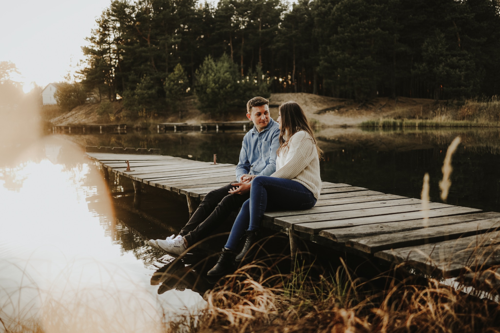 Para siedząca na drewnianym molo nad jeziorem, w tle las iglasty i zachodzące słońce, tworzące romantyczną atmosferę.