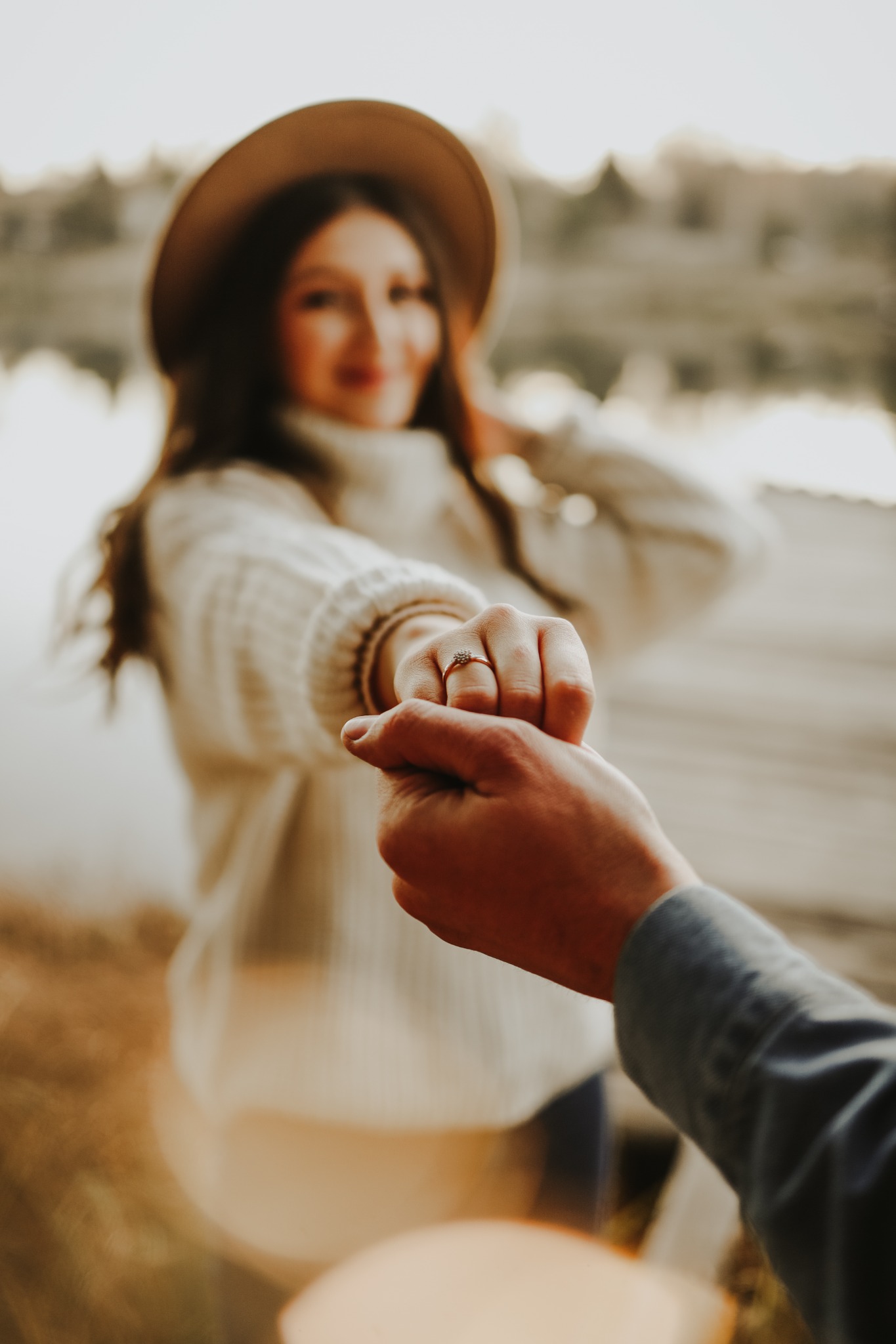 Para trzymająca się za ręce, pierścionek zaręczynowy na palcu kobiety, ona w beżowym swetrze i kapeluszu, on w jeansowej koszuli, rozmyte tło jeziora i drzew.