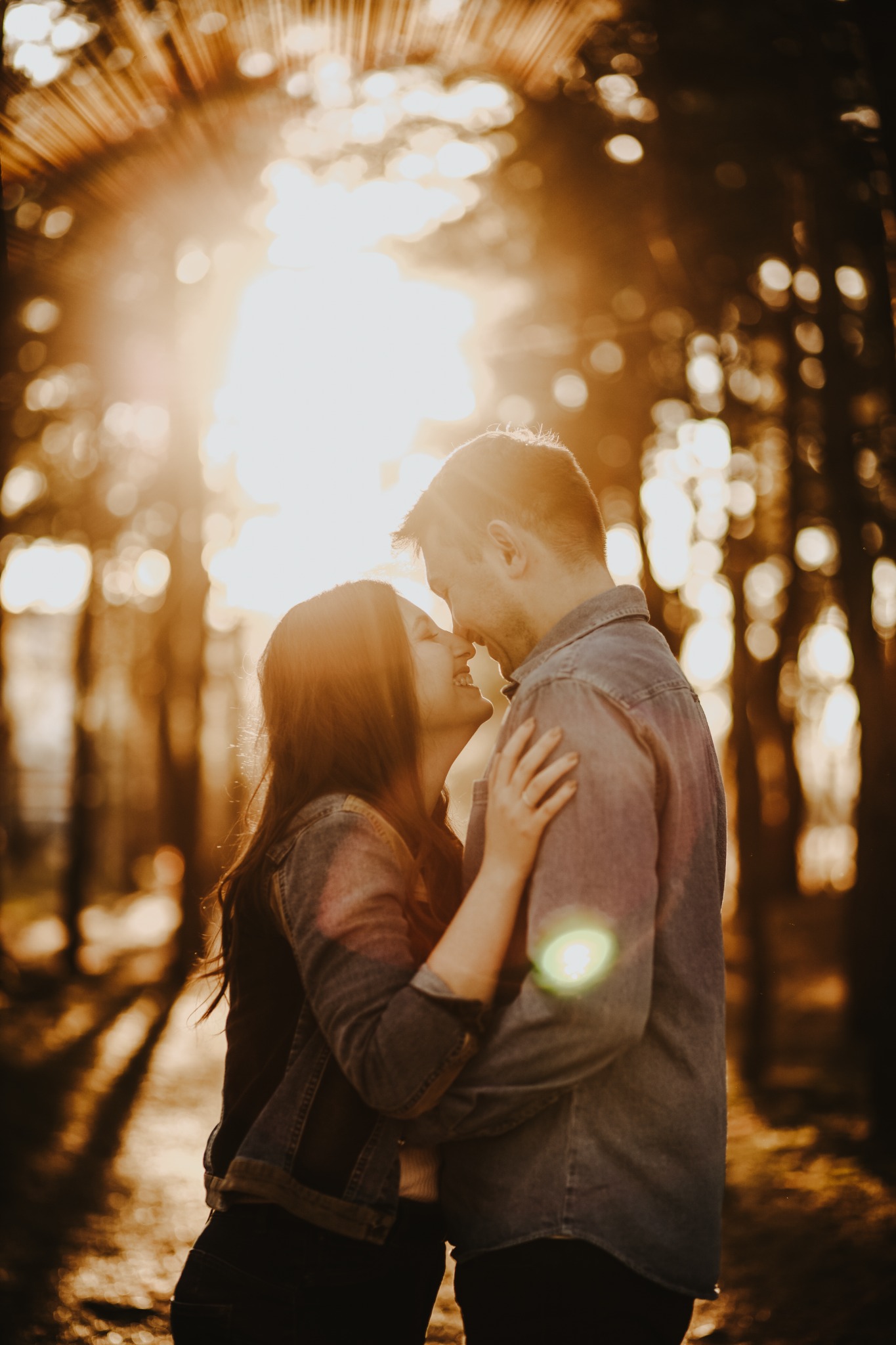 Zakochana para w objęciach na tle drzew i zachodzącego słońca, delikatne promienie oświetlają ich twarze.