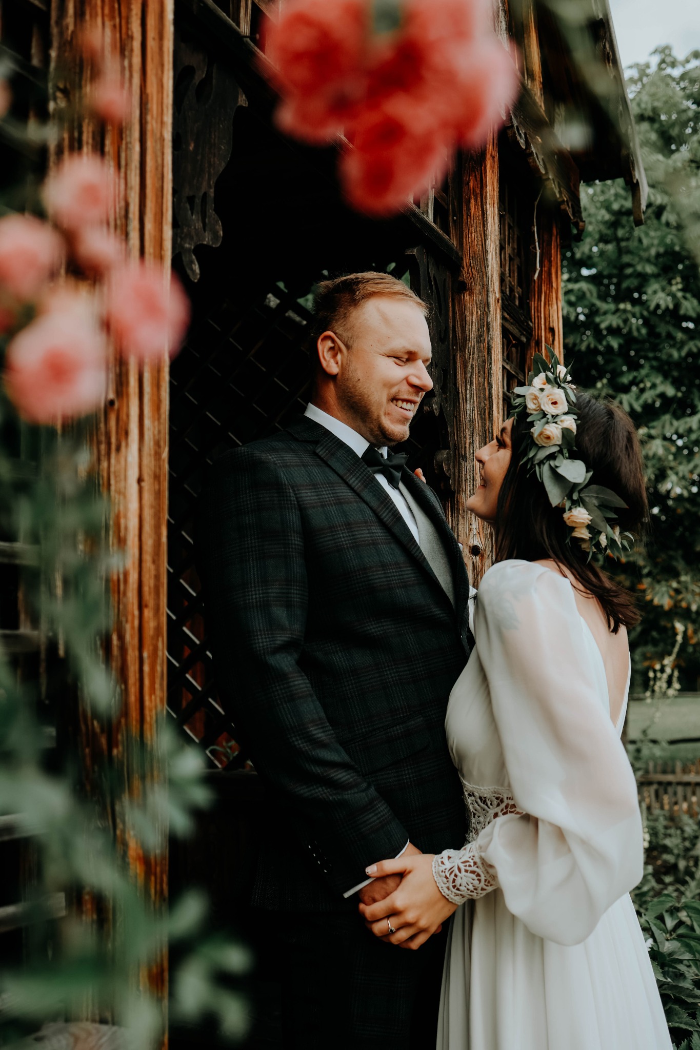 Uśmiechnięta para młoda, panna młoda z wiankiem z białych róż i zielonych liści, pan młody w ciemnej kraciastej marynarce i muszce, trzymają się za ręce na tle drewnianej altanki i zieleni.