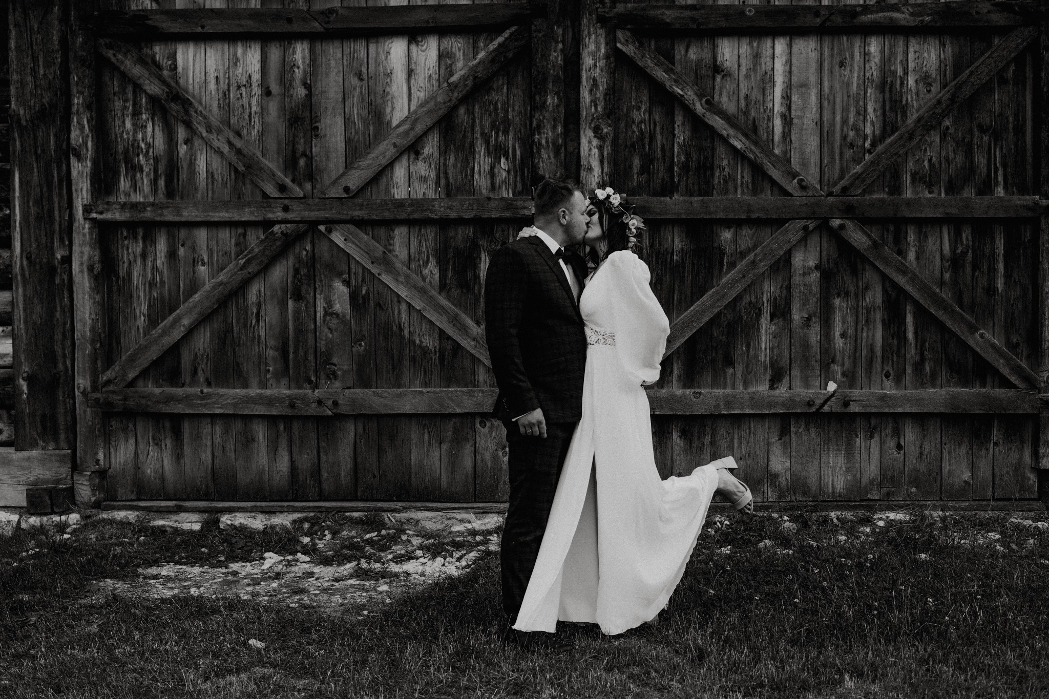 Czarno-białe zdjęcie całującej się pary młodej na tle rustykalnych drewnianych wrót stodoły, panna młoda w sukni z rozcięciem i wiankiem na głowie, uniesiona noga w bucie.