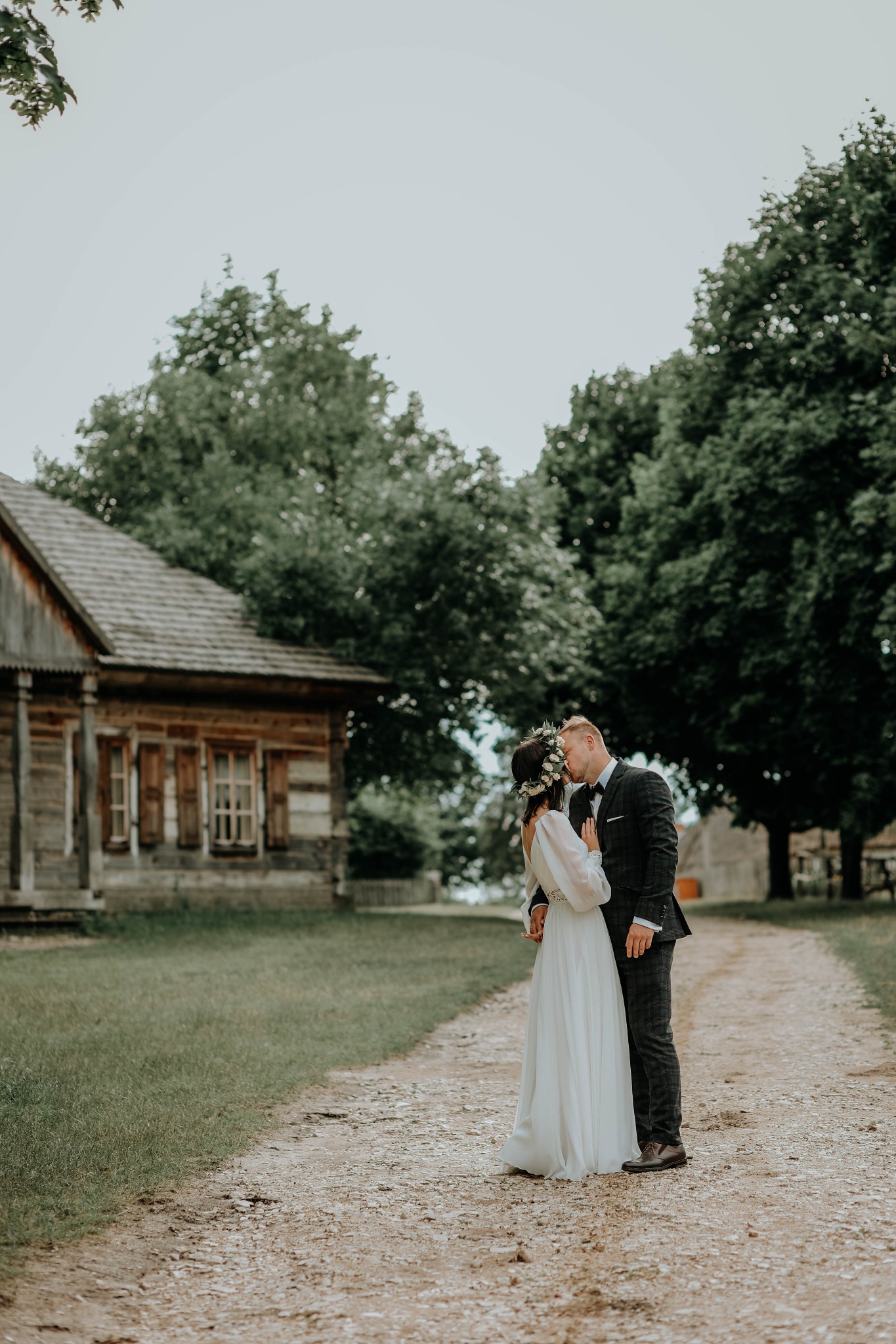 Para młoda całuje się na wiejskiej drodze, obok drewnianego domu z brązowymi okiennicami, otoczona zielenią drzew.