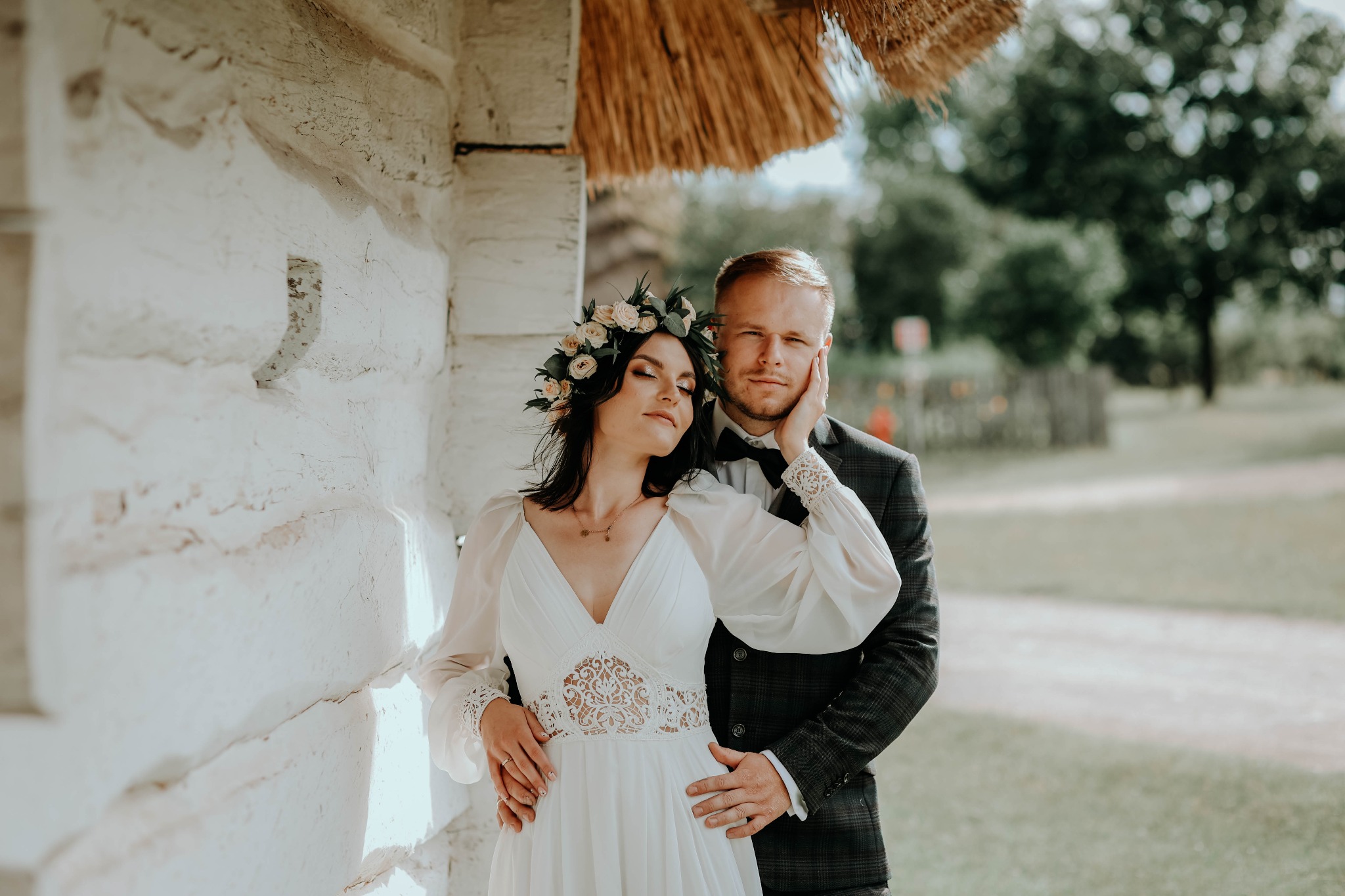 Para młoda w rustykalnej sesji ślubnej; panna młoda w białej sukni z koronkowym detalem i wiankiem z jasnych kwiatów, pan młody w ciemnym garniturze w kratę, pozują na tle białej ściany budynku...