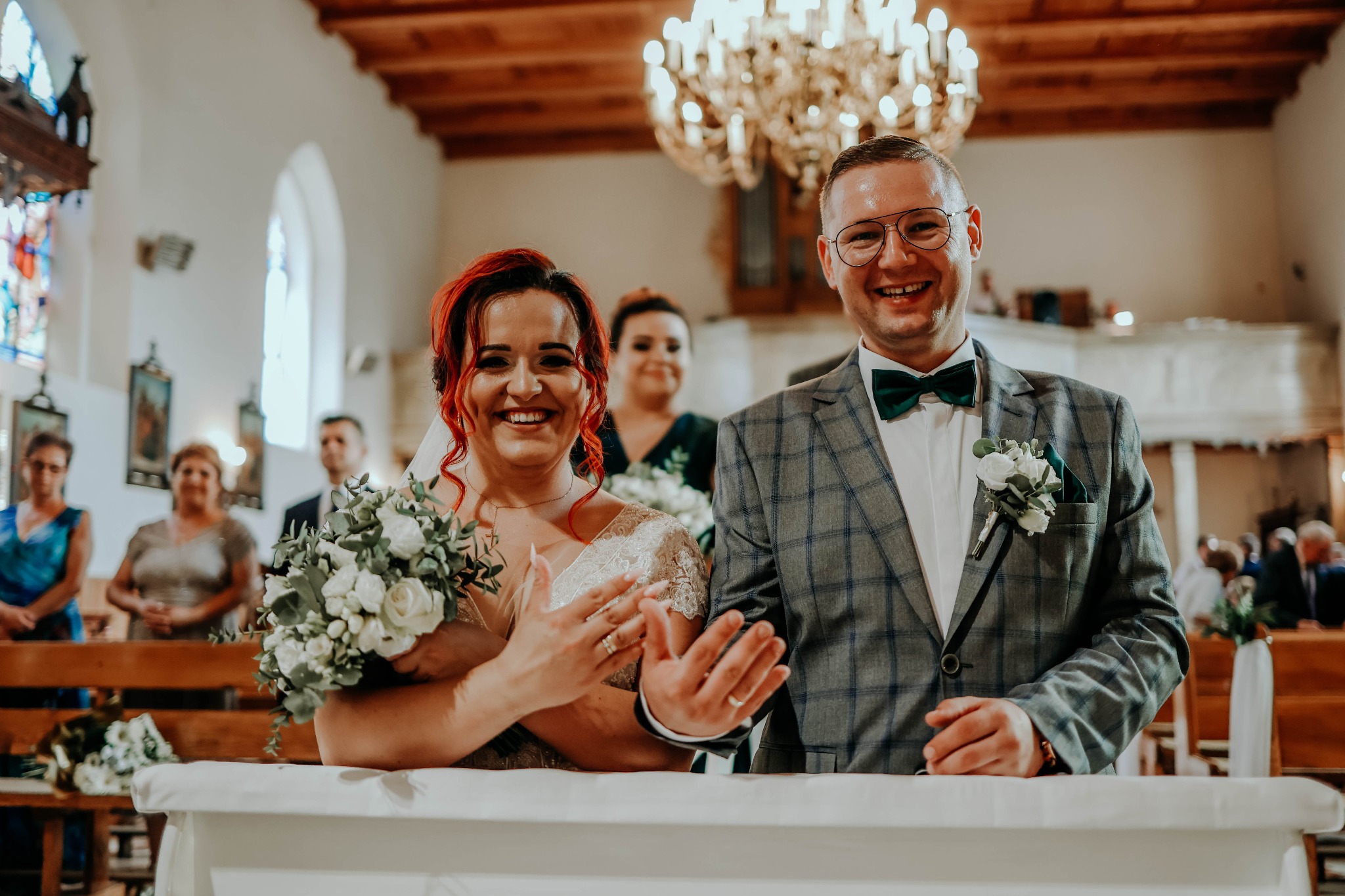Uśmiechnięta para młoda w kościele, panna młoda z rudymi włosami trzyma bukiet, pan młody w szarej marynarce w kratę i zielonej muszce, w tle goście i żyrandol.