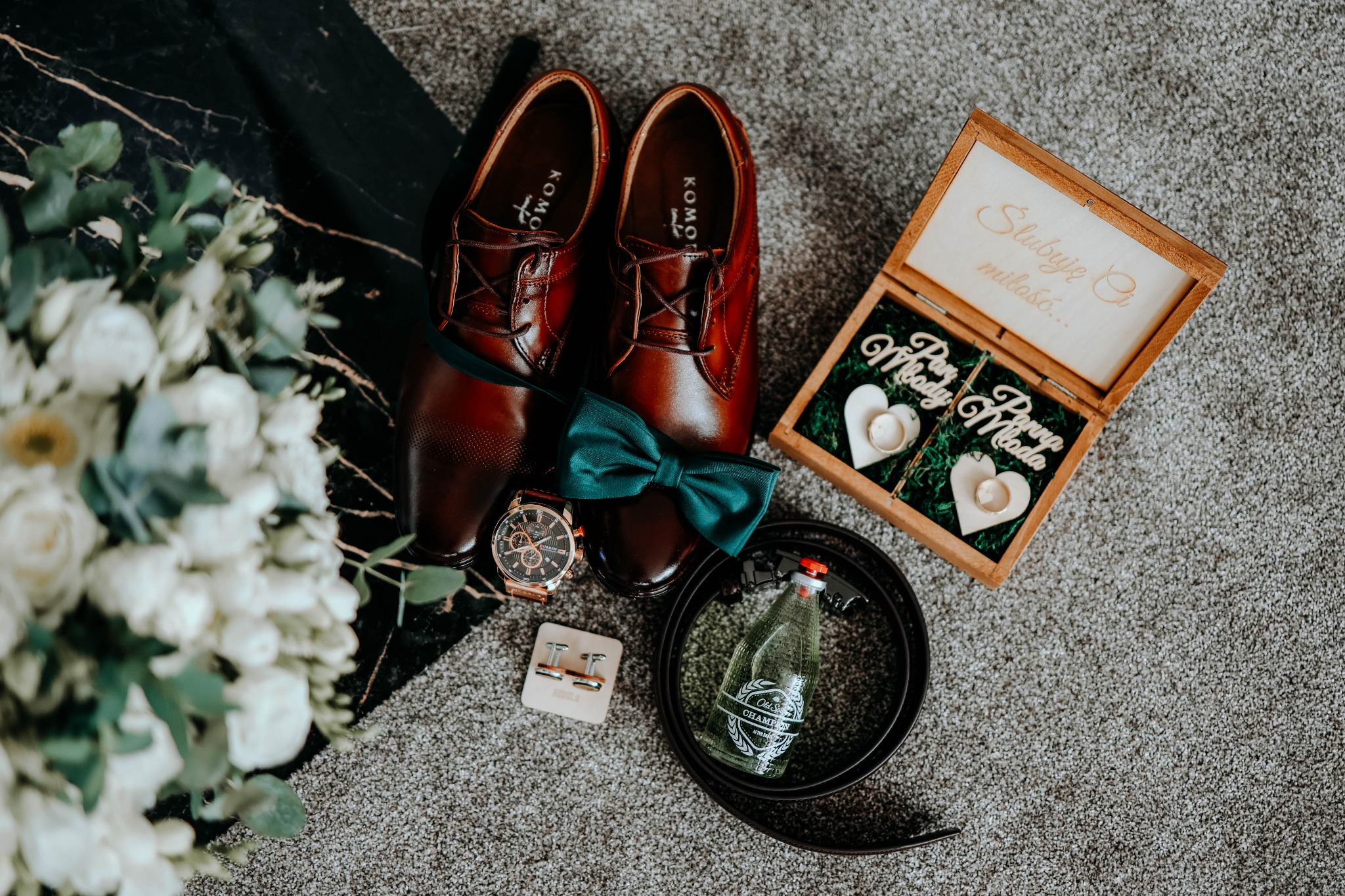 Brązowe skórzane buty, zielona mucha, zegarek, spinki do mankietów, pasek, miniaturowa butelka szampana i drewniane pudełko na obrączki z napisem 'Ślubuję Ci miłość...' oraz 'Pan Młody' i 'Panna...