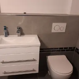 Nowoczesna łazienka w szarościach z wiszącą szafką umywalkową z trzema szufladami, chromowaną armaturą, toaletą i dekoracyjną listwą z ciemnych płytek.