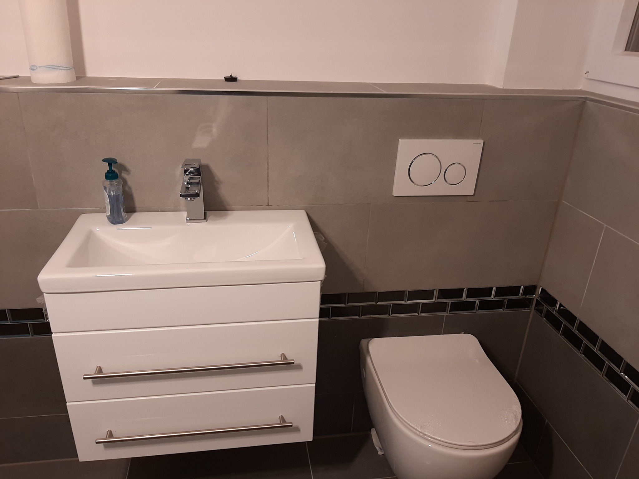 Nowoczesna łazienka w szarościach z wiszącą szafką umywalkową z trzema szufladami, chromowaną armaturą, toaletą i dekoracyjną listwą z ciemnych płytek.