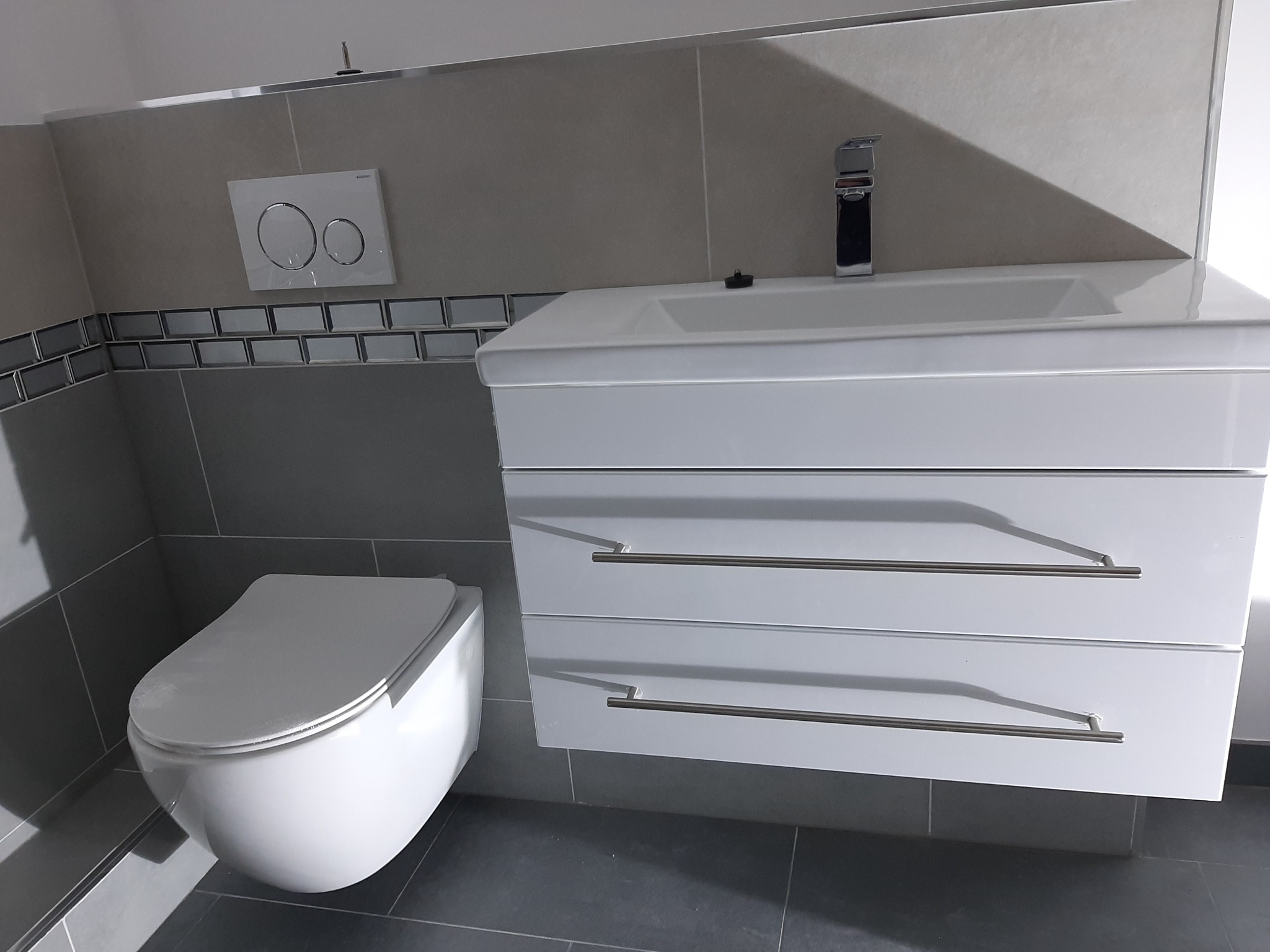 Nowoczesna, minimalistyczna łazienka z podwieszaną toaletą, białą szafką z umywalką i chromowaną baterią oraz szarymi płytkami.