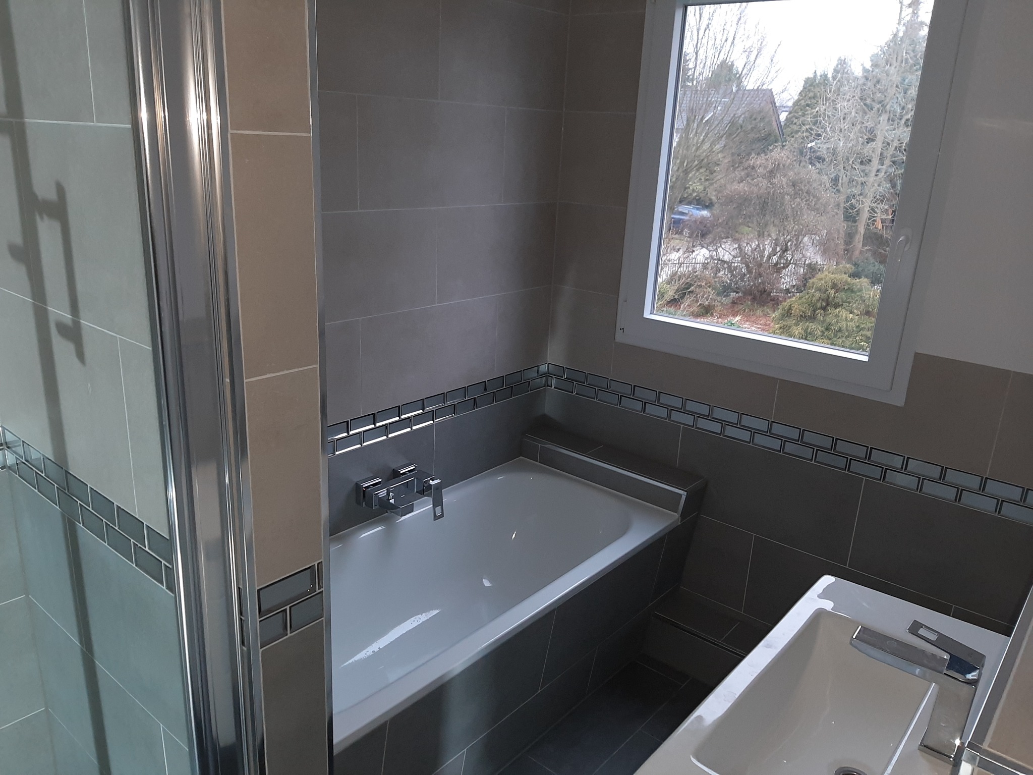 Nowoczesna łazienka z szarymi płytkami, mozaiką i białymi elementami armatury, widok na ogród przez okno, kabina prysznicowa z chromowanymi profilami.