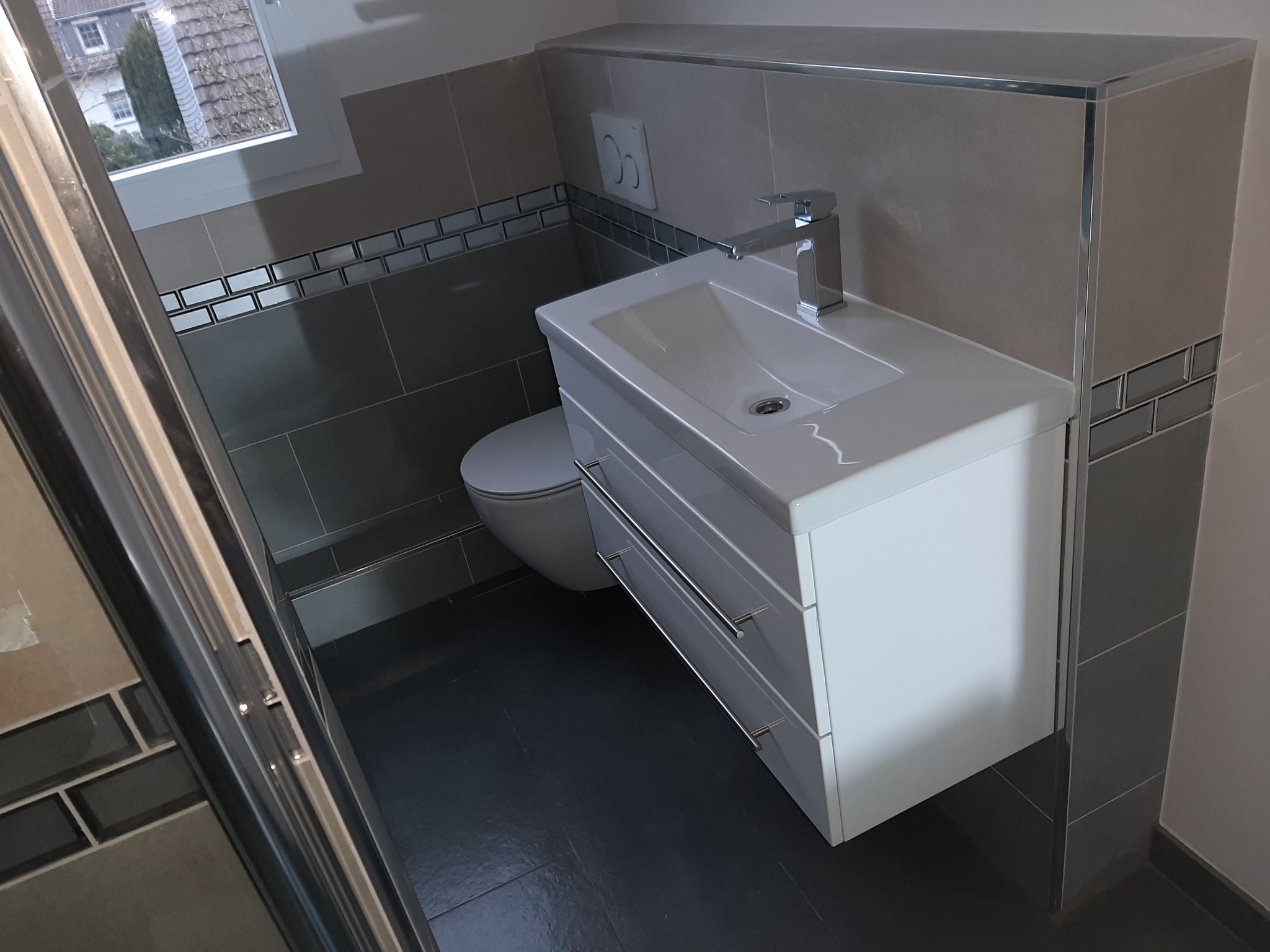 Nowoczesna łazienka po remoncie: biała szafka pod umywalką z trzema szufladami i chromowaną baterią, wisząca toaleta, szare płytki z dekoracyjnym pasem mozaiki, okno z widokiem na dom.