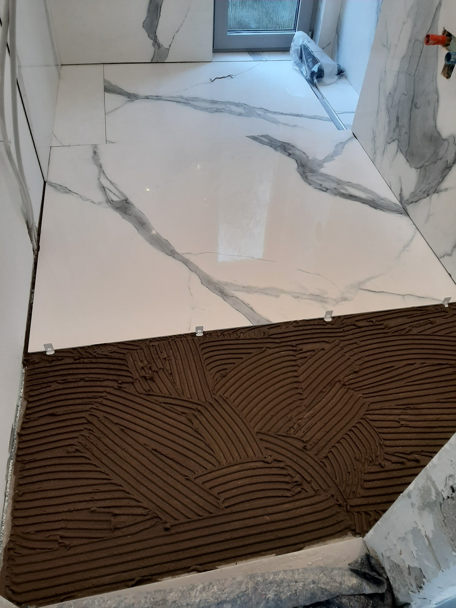 Układanie białych płytek imitujących marmur na podłodze w łazience, widoczny klej i plastikowe krzyżyki dystansowe.