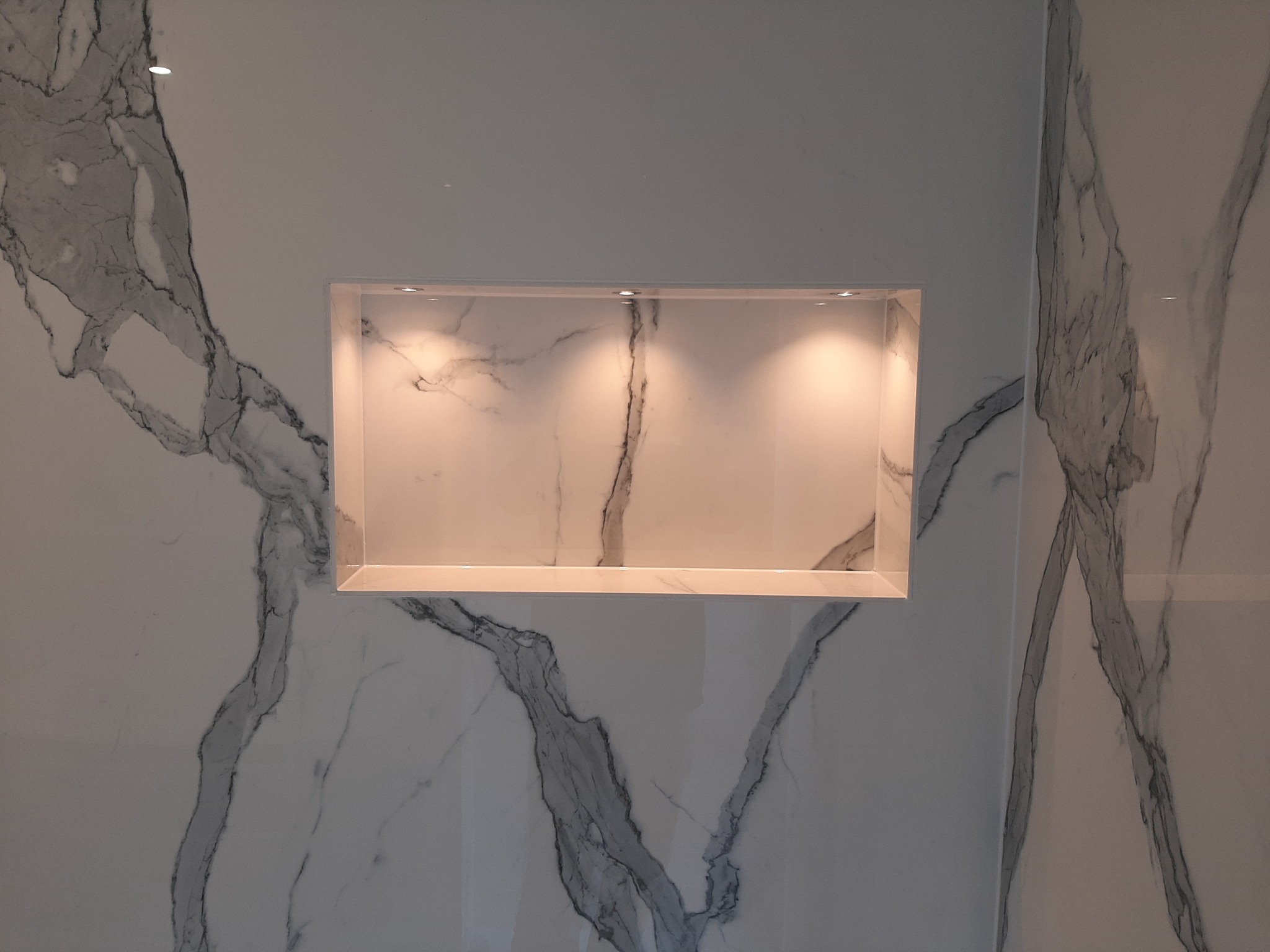 Wnęka z podświetleniem LED w ścianie wyłożonej płytami imitującymi biały marmur z szarymi żyłami.