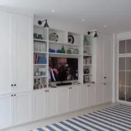 Biała zabudowa meblowa na ścianie z półkami, szafkami i miejscem na telewizor, uzupełniona oświetleniem punktowym i dekoracjami.