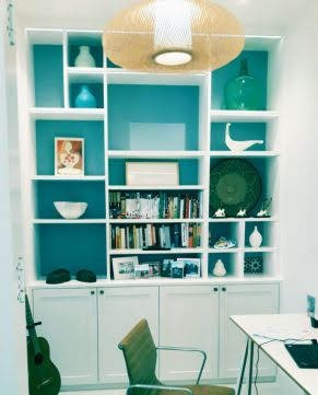 Biały regał wnękowy z turkusowym tłem półek, wypełniony dekoracjami i książkami, oświetlony ozdobną lampą wiszącą, z gitarą opartą o bok i krzesłem na pierwszym planie.