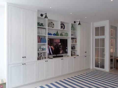 Biała zabudowa meblowa na ścianie z półkami, szafkami i miejscem na telewizor, uzupełniona oświetleniem punktowym i dekoracjami.