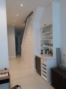 Białe wnętrze domu z zabudową pod schodami, obejmującą szafki, półki z dekoracyjnym szkłem i małą lodówką. Schody z ciemną poręczą i białymi tralkami. W tle widoczny korytarz z oświetleniem...