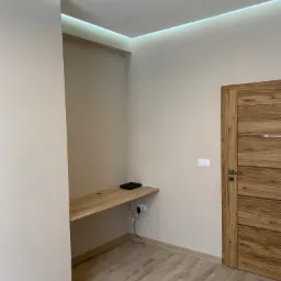 Wnętrze pokoju po remoncie: drewnopodobna podłoga, beżowe ściany, drewniane drzwi z poziomymi intarsjami, podwieszany drewniany blat biurka z czarnym routerem, oświetlenie LED pod sufitem.