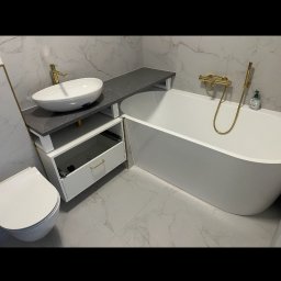 Gastro Bar Service Adam Hołoga - Nowoczesna łazienka z wolnostojącą wanną, umywalką nablatową na szarym blacie i złotą armaturą, wykończona płytkami imitującymi marmur.