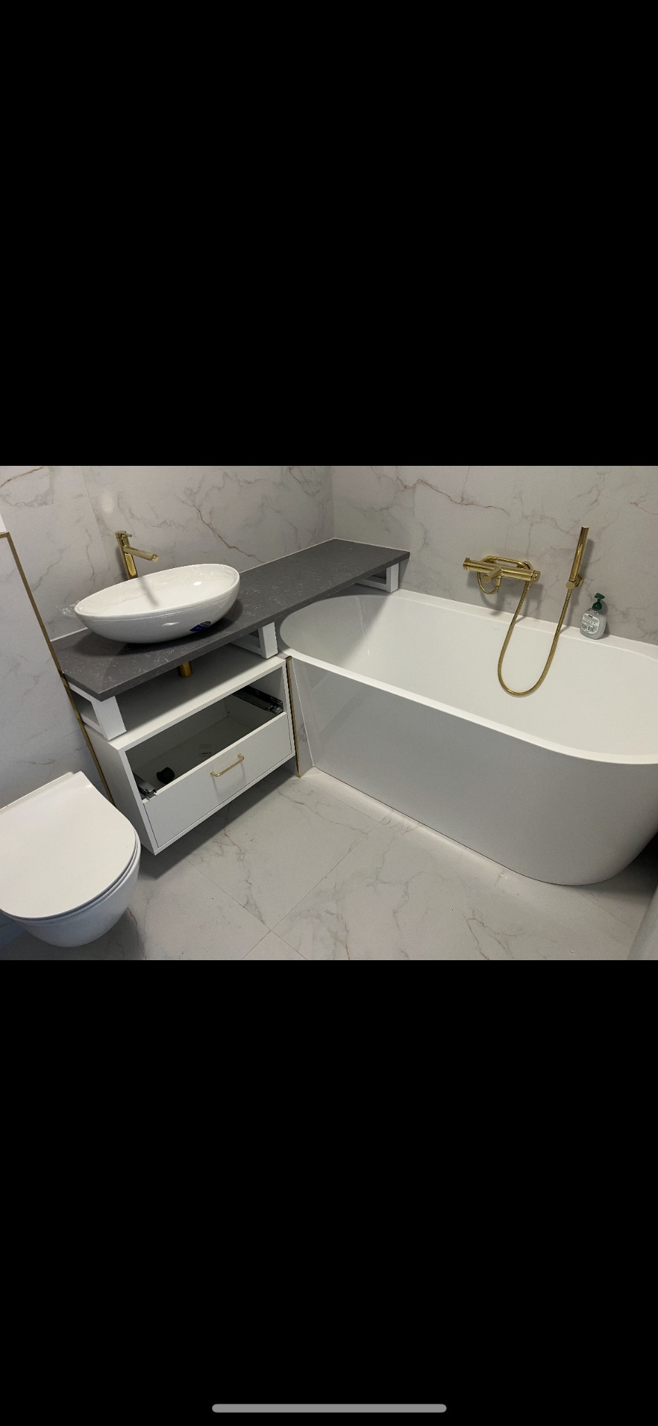 Nowoczesna łazienka z białą armaturą, umywalką nablatową, wanną wolnostojącą i złotą baterią, wykończona płytkami imitującymi marmur.