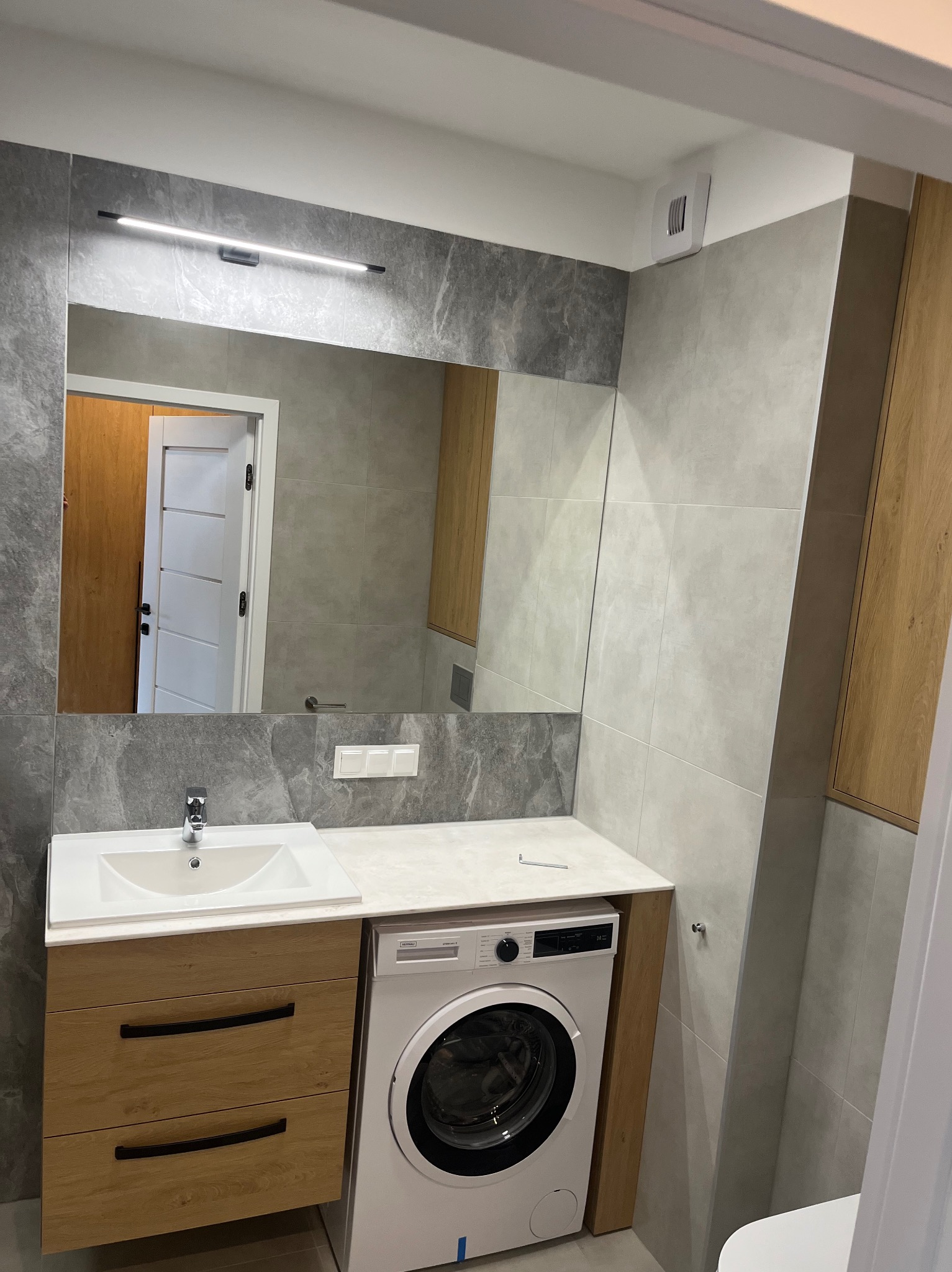 Nowoczesna łazienka po remoncie: szare płytki imitujące marmur, biała umywalka nablatowa na szafce z drewnopodobnymi frontami z czarnymi uchwytami, pralka pod blatem, duże lustro z oświetleniem LED...