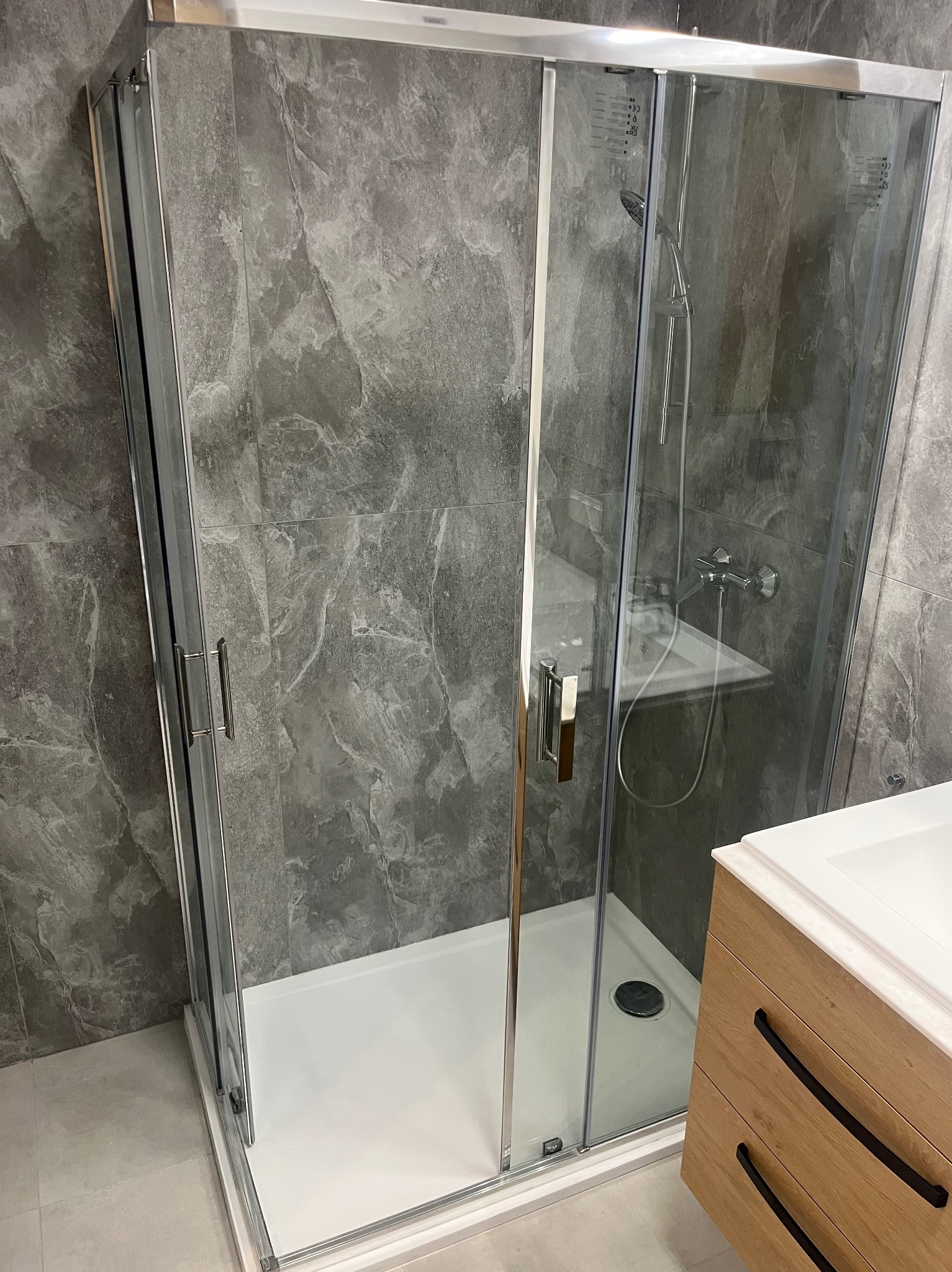 Kabina prysznicowa z przesuwnymi drzwiami, chromowanymi profilami i brodzikiem, na tle ściany wyłożonej szarymi płytkami imitującymi kamień. Widoczna armatura i fragment szafki pod umywalkę...