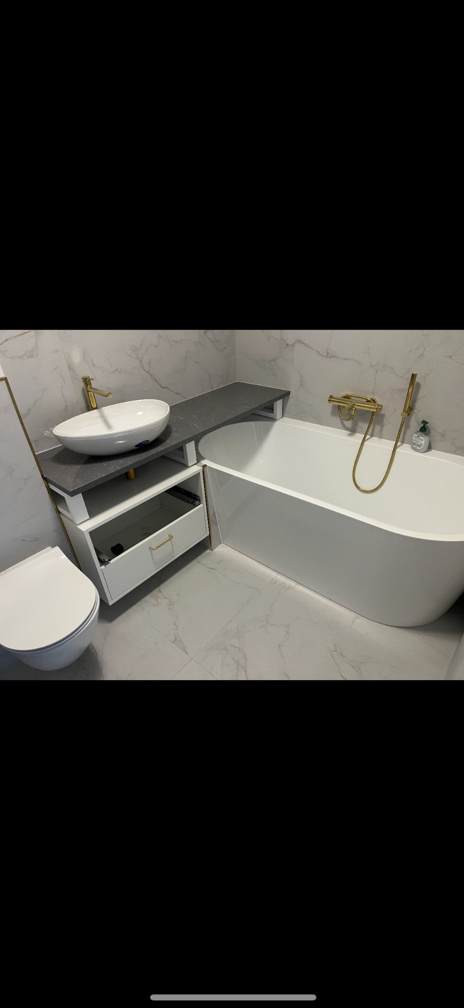 Nowoczesna łazienka z wolnostojącą wanną, umywalką nablatową na szarym blacie i złotą armaturą, wykończona płytkami imitującymi marmur.