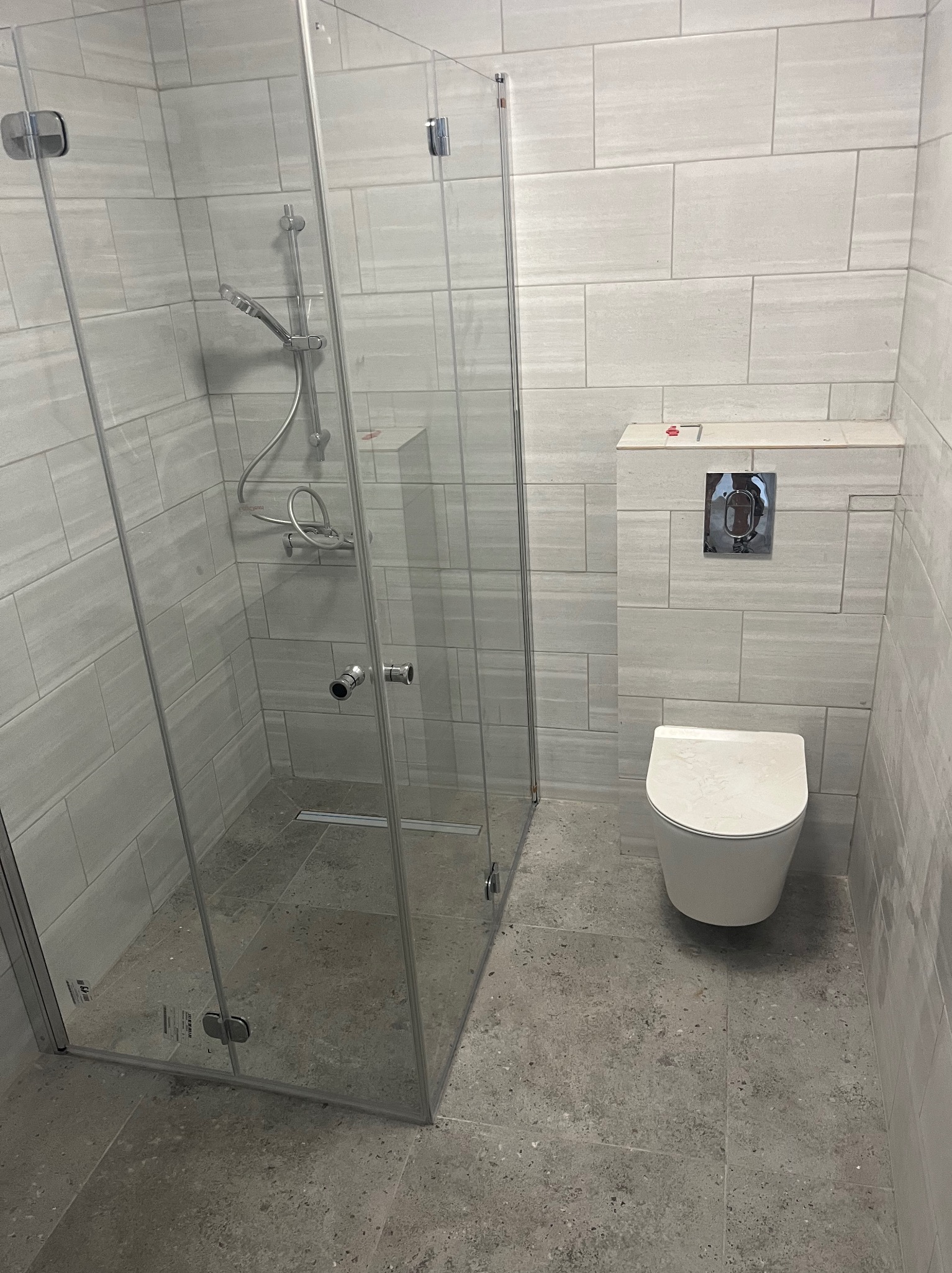 Nowoczesna łazienka z przeszkloną kabiną prysznicową i podwieszaną toaletą, wykończona płytkami w odcieniach szarości z delikatnym wzorem.