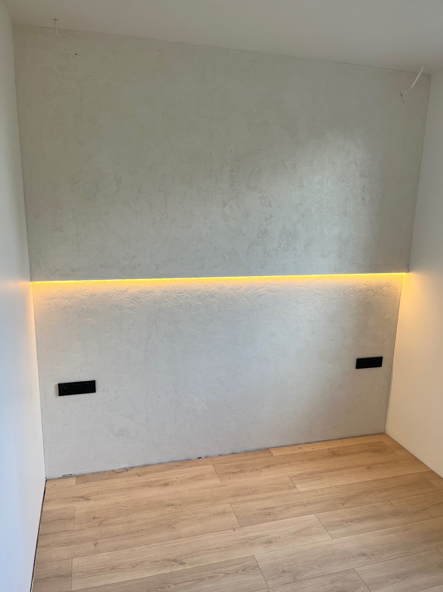 Ściana z dekoracyjną fakturą i podświetleniem LED, widoczne gniazdka elektryczne i panele podłogowe w jasnym odcieniu drewna.