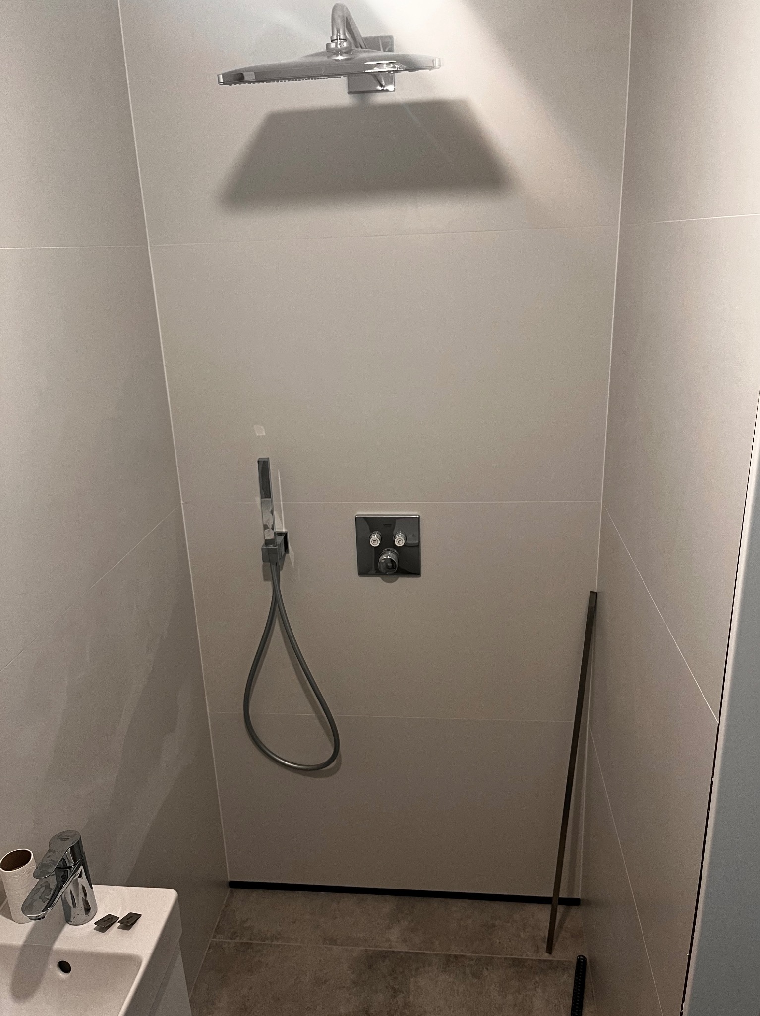 Nowoczesna kabina prysznicowa z deszczownicą i baterią podtynkową, wykończona płytkami w odcieniach szarości, widoczna umywalka z baterią i akcesoriami.