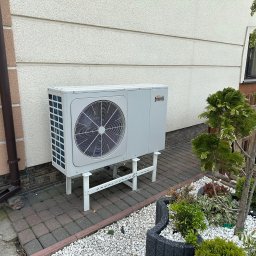 VIA-SYSTEM - Biała jednostka zewnętrzna z logo Ferroli, zamontowana na metalowym stelażu przy ścianie budynku z ozdobnym ogrodem skalnym obok.