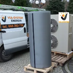 Biały samochód dostawczy z otwartymi drzwiami i reklamą firmy VIA-SYSTEM Energia, obok szary zbiornik buforowy na palecie oraz jednostki zewnętrzne klimatyzacji również na paletach, ustawione...