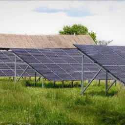 Rząd paneli słonecznych zamontowanych na metalowych konstrukcjach na tle wiejskiego krajobrazu z budynkami gospodarczymi i silosami.