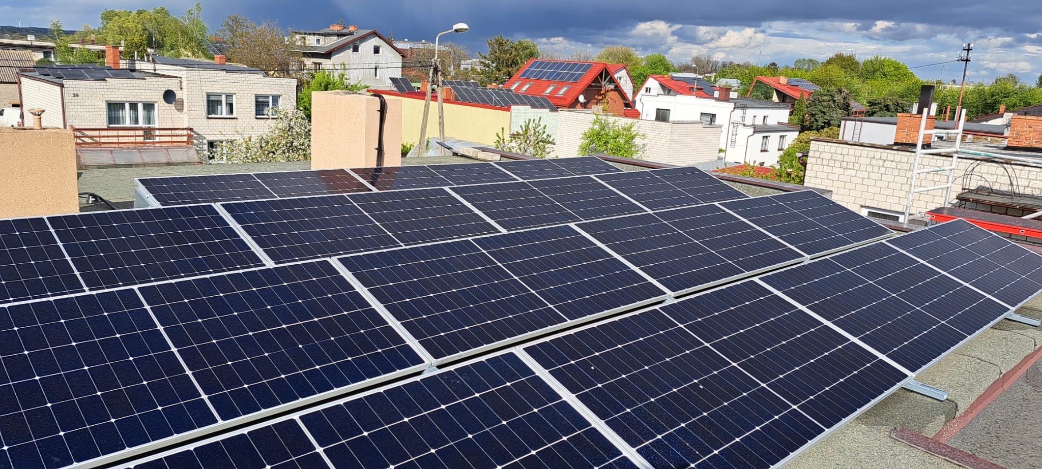 Rząd paneli słonecznych na płaskim dachu budynku mieszkalnego, z widokiem na inne domy i pochmurne niebo.
