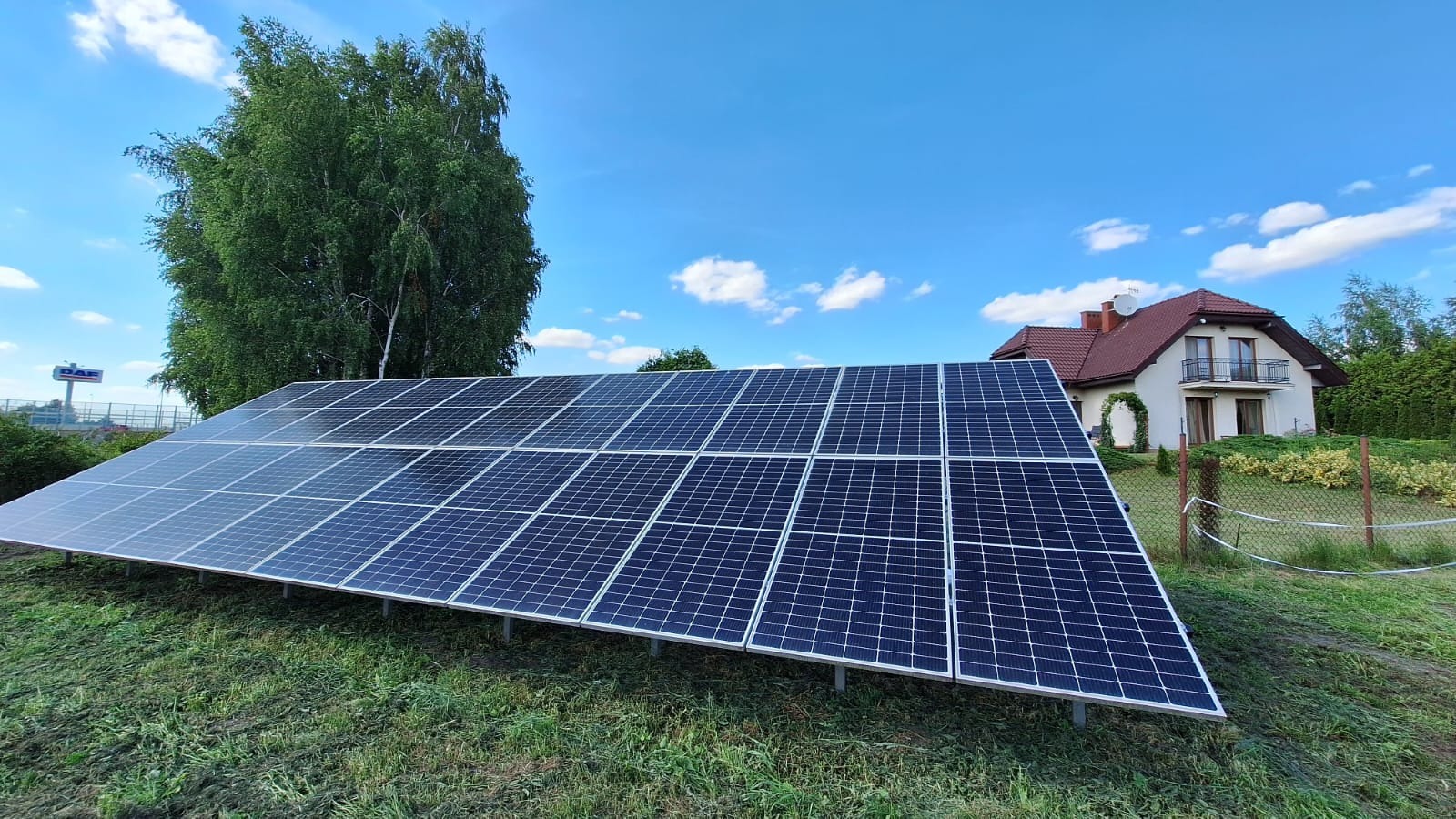 Rząd paneli słonecznych na trawiastym zboczu przed domem jednorodzinnym z czerwonym dachem, błękitne niebo w tle.