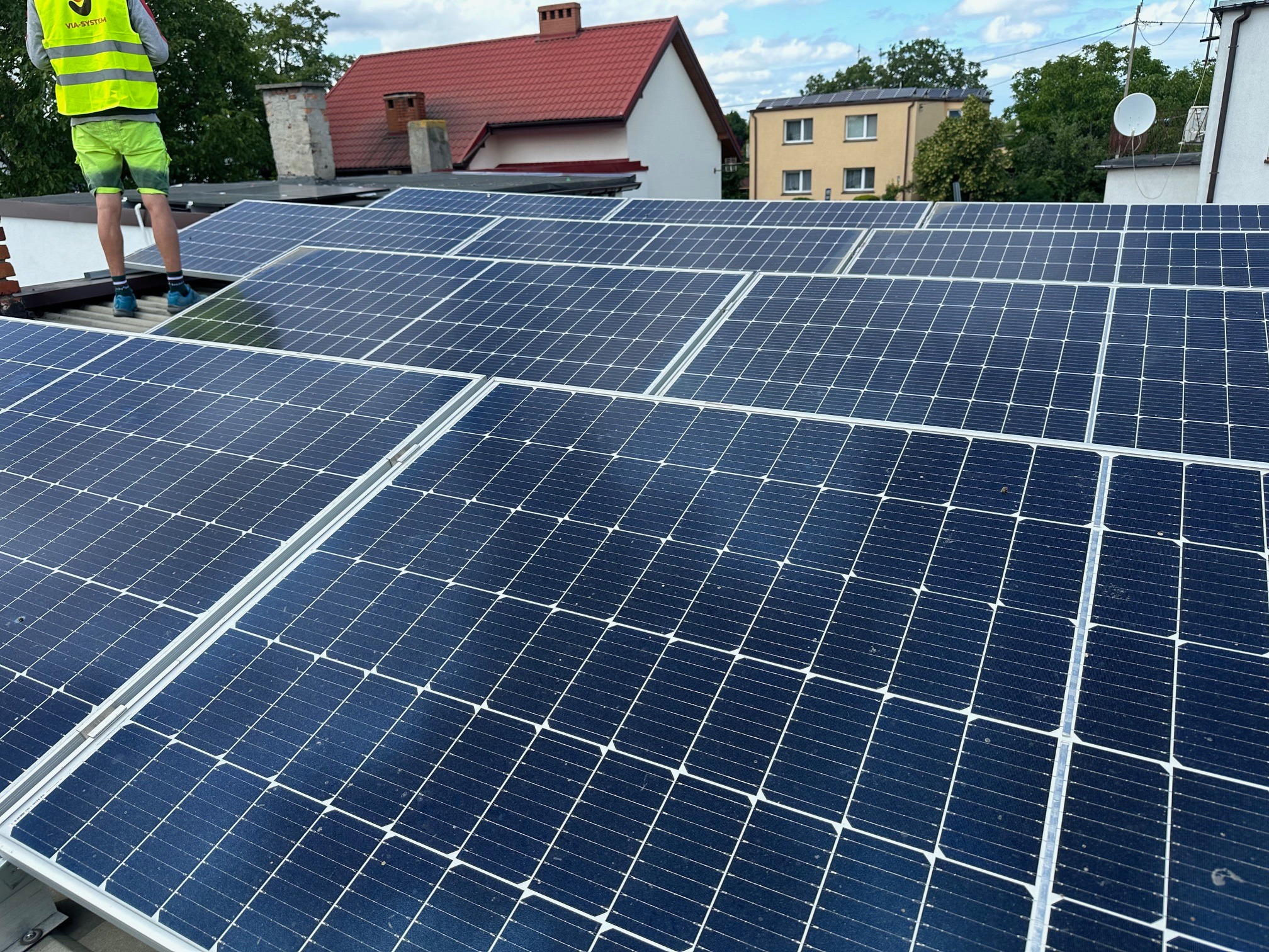 Instalacja paneli fotowoltaicznych na dachu domu jednorodzinnego z widocznym fragmentem pracownika w odblaskowej kamizelce i neonowych spodenkach.