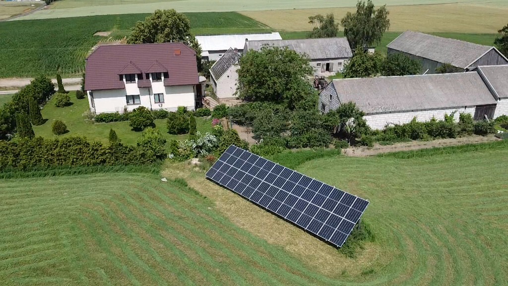 Panel słoneczny zainstalowany na trawniku przed wiejskim domem z budynkami gospodarczymi w tle, widok z góry.