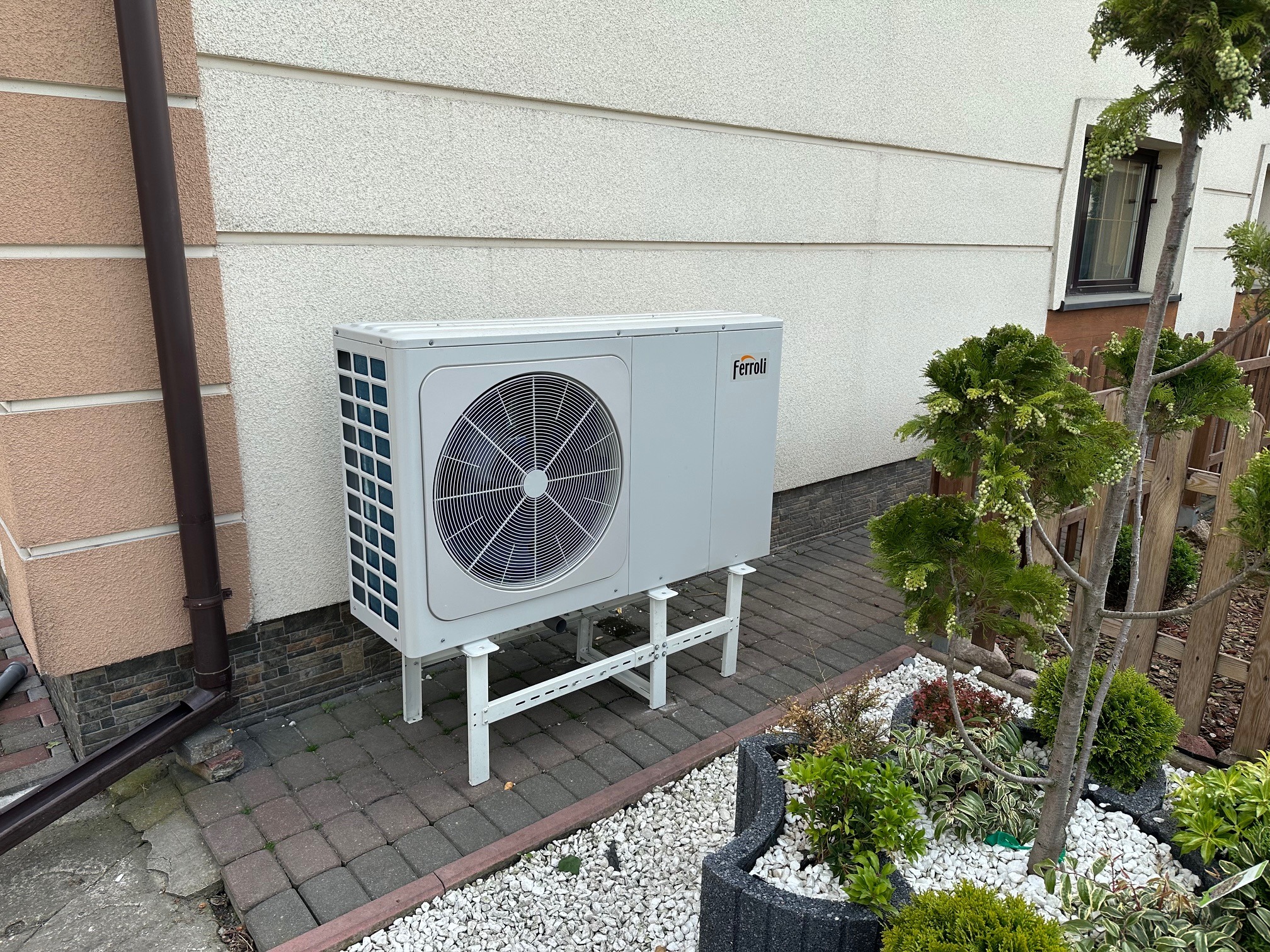 Biała jednostka zewnętrzna z logo Ferroli, zamontowana na metalowym stelażu przy ścianie budynku z ozdobnym ogrodem skalnym obok.