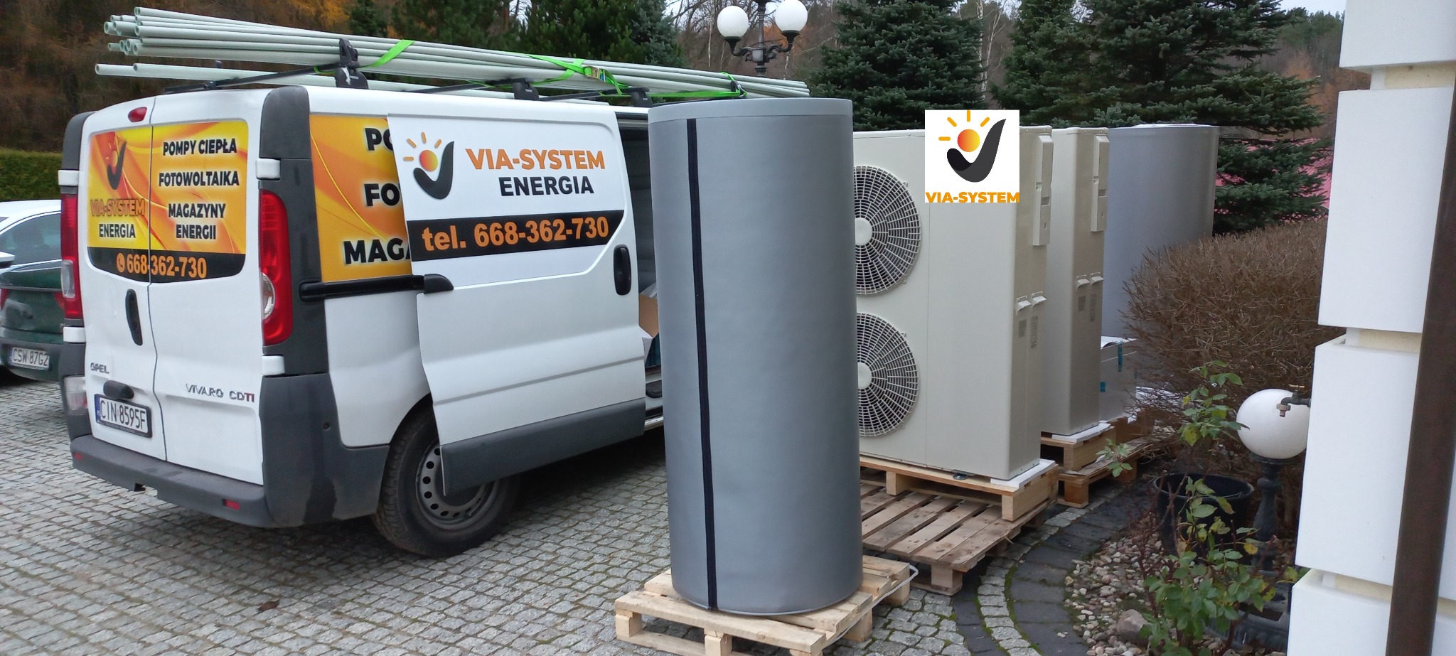 Biały samochód dostawczy z otwartymi drzwiami i reklamą firmy VIA-SYSTEM Energia, obok szary zbiornik buforowy na palecie oraz jednostki zewnętrzne klimatyzacji również na paletach, ustawione...