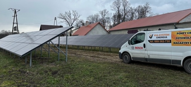 Rząd paneli fotowoltaicznych na metalowej konstrukcji w wiejskim otoczeniu, z zaparkowanym białym vanem firmy Via-System Energia z reklamą pomp ciepła, fotowoltaiki i magazynów energii.