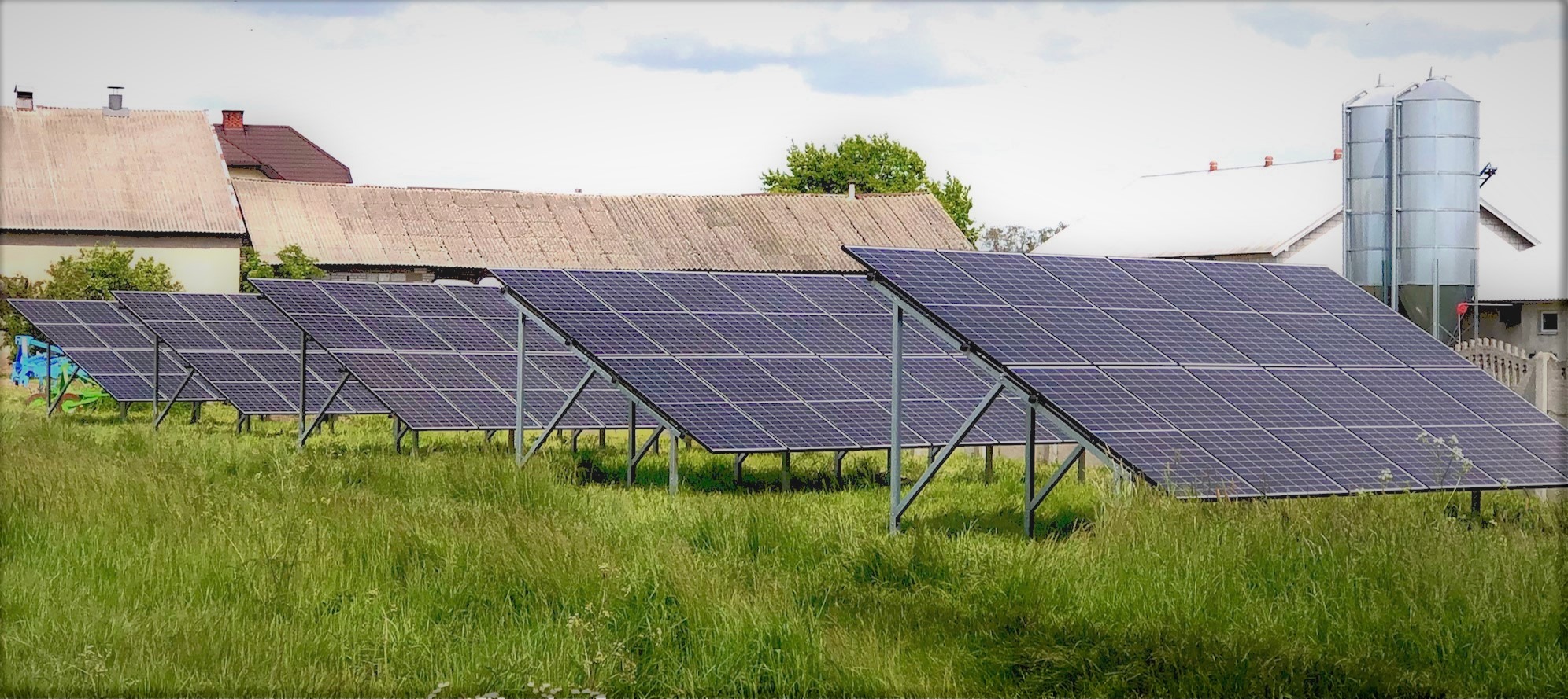 Rząd paneli słonecznych zamontowanych na metalowych konstrukcjach na tle wiejskiego krajobrazu z budynkami gospodarczymi i silosami.