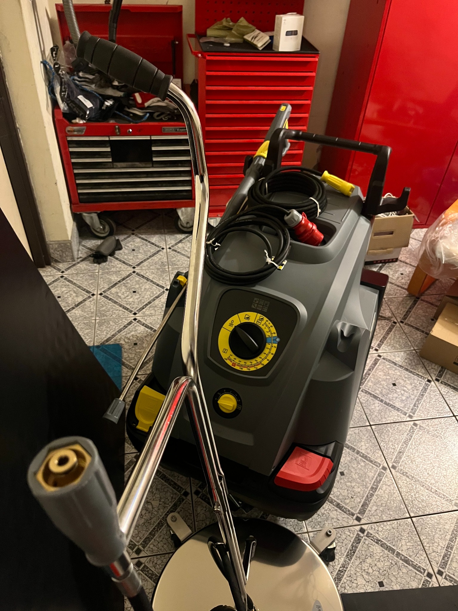 Wysokociśnieniowa myjka Karcher z akcesoriami, w tle czerwone szafki warsztatowe i wzorzysta podłoga.