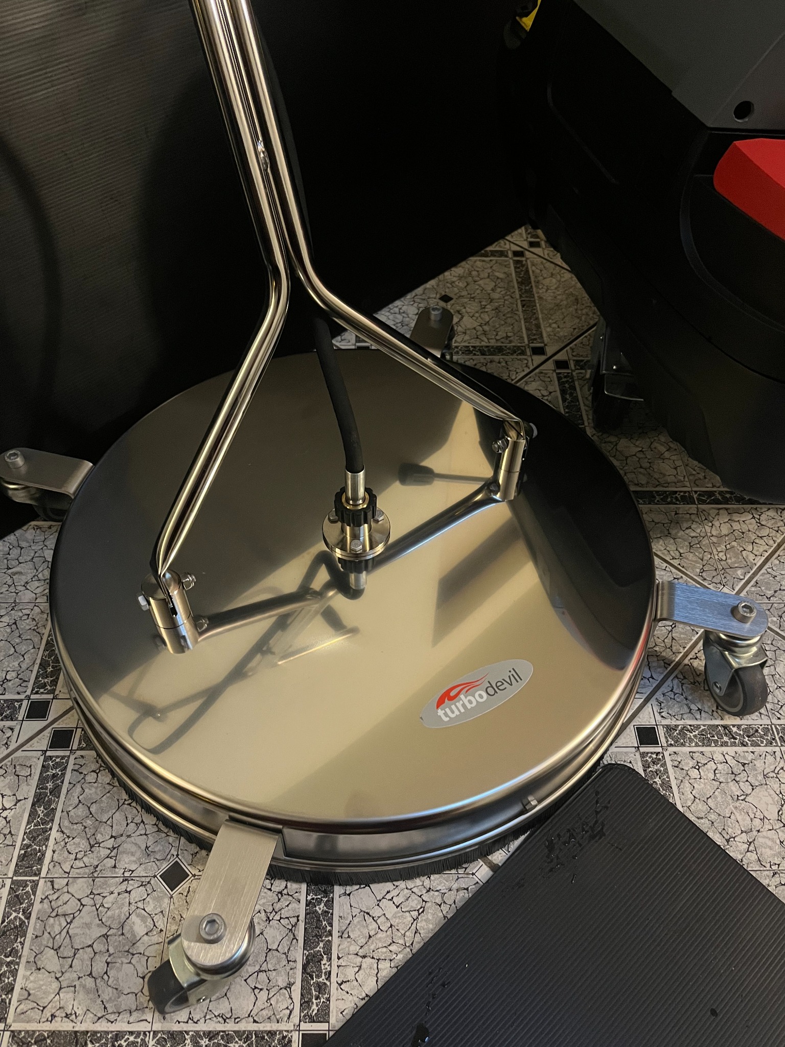 Okrągła, metalowa myjka ciśnieniowa do powierzchni płaskich marki Turbodevil z dyszami pod spodem, widoczna na tle płytek podłogowych.